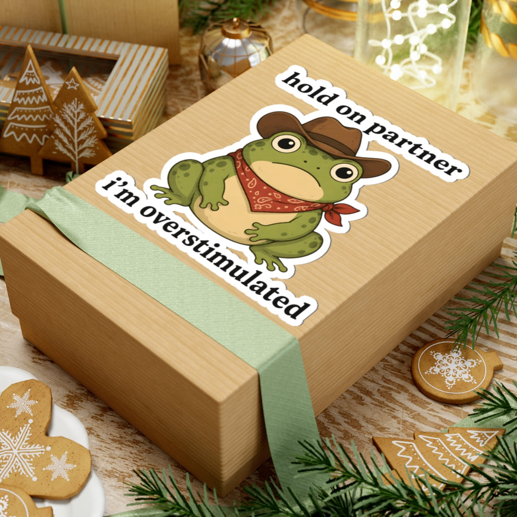 A wooden gift box wrapped with a pale green ribbon, featuring a cartoon frog sticker that says 'hold on partner i’m overstimulated.' The box is styled on a tabletop with holiday cookies, evergreen branches, and warm decorative lights in the background.
