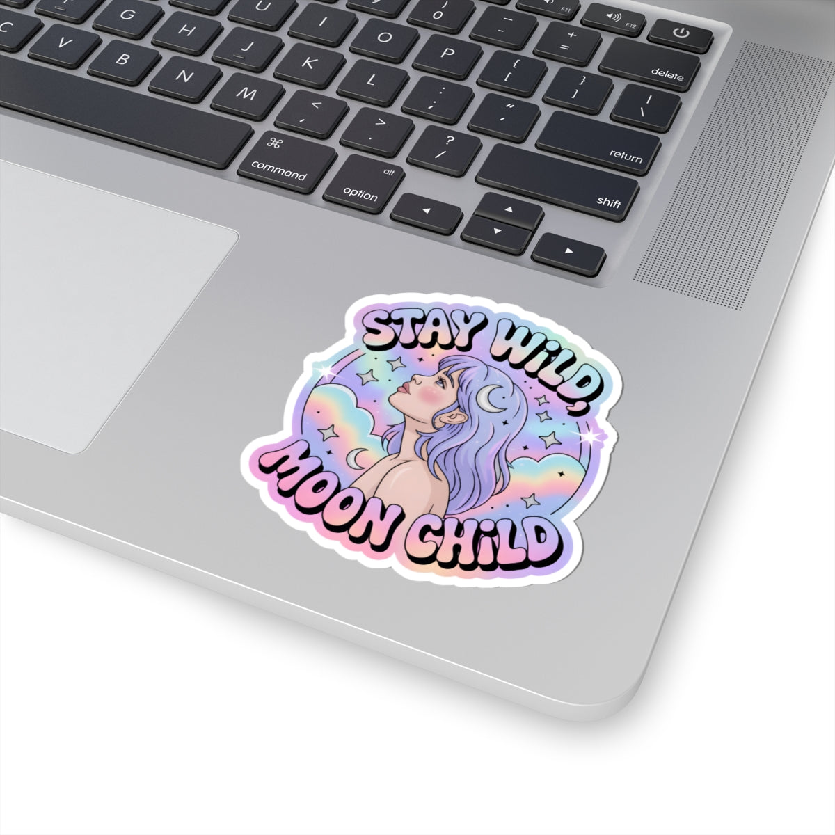 A kiss-cut vinyl sticker with the text 'STAY WILD MOON CHILD' and an illustration of a pastel-haired fantasy girl, shown applied to the palm rest of a silver laptop.