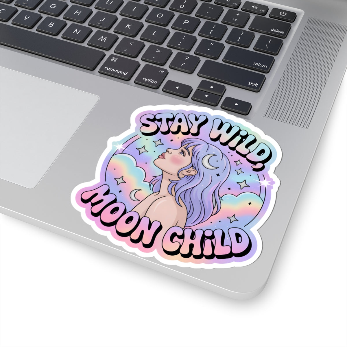 A kiss-cut vinyl sticker with a pastel rainbow illustration of a girl looking up at a crescent moon, with the text 'STAY WILD, MOON CHILD' in bubbly outlined lettering. The sticker is shown applied to the corner of a silver laptop keyboard.