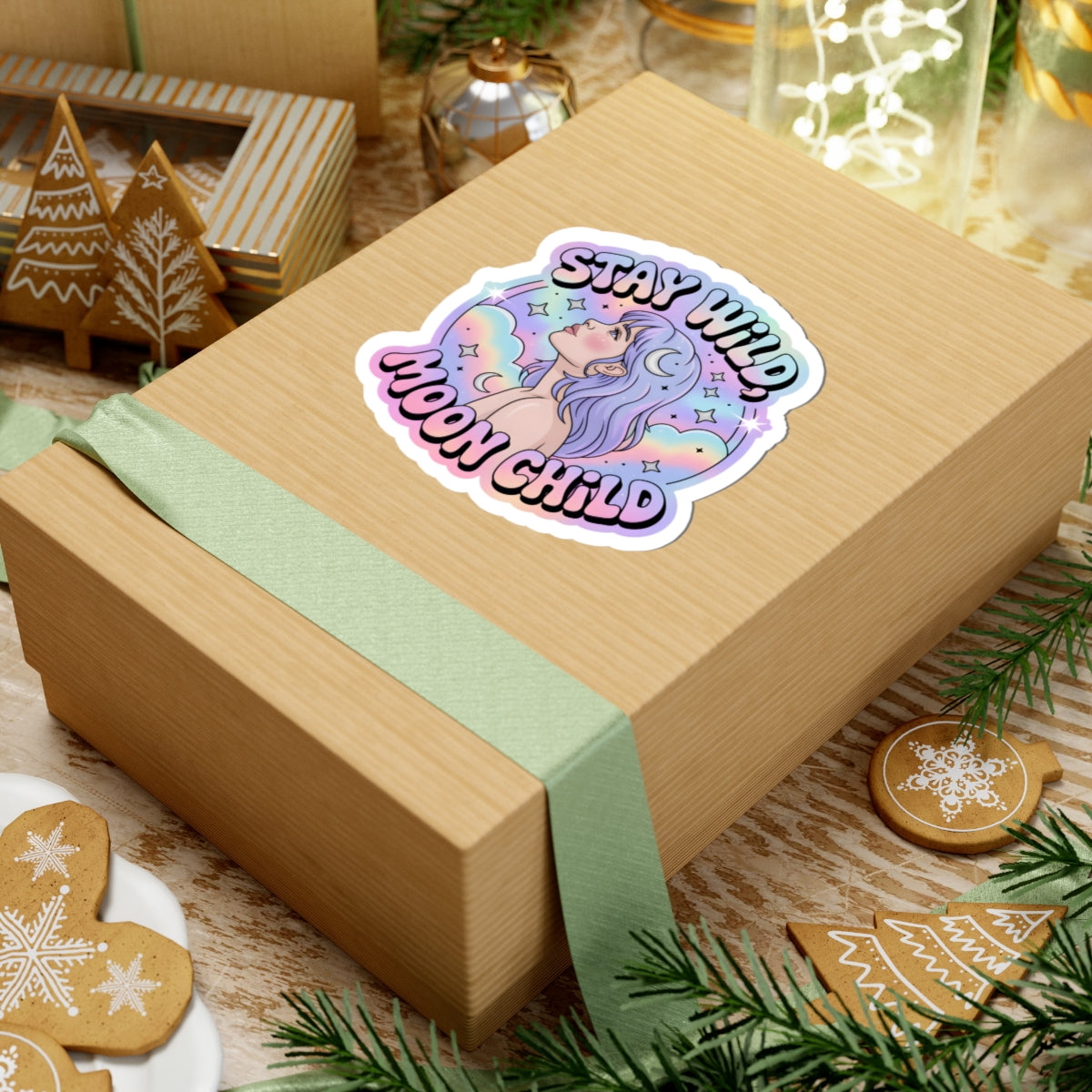 A kraft paper gift box tied with a pale green ribbon, featuring a colorful die-cut sticker that reads 'STAY WILD MOON CHILD' with an illustrated moon character, styled on a tabletop with holiday decorations.