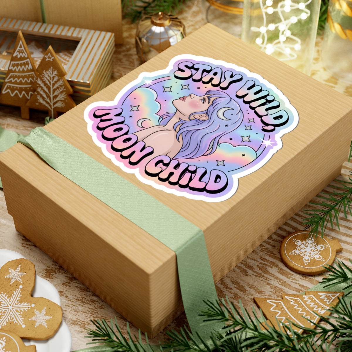 A kiss-cut sticker with an anime-style moon goddess illustration and the text 'STAY WILD, MOON CHILD' in pink lettering, shown placed on top of a kraft gift box wrapped with a pale green ribbon. The scene is styled with holiday cookies, pine branches, and warm ambient lights.