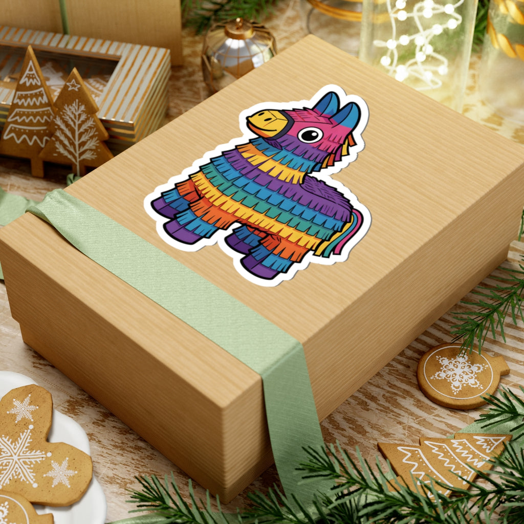 A rectangular kraft paper gift box wrapped with a pale green ribbon, topped with a colorful piñata-shaped sticker. The box is styled on a tabletop with holiday cookies, evergreen branches, and other gift items in the background.