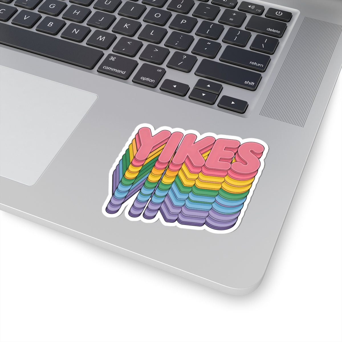 A kiss-cut vinyl sticker with the word 'YIKES' in pink 3D-style lettering over a rainbow-striped wavy shape, shown applied to the lower part of a silver laptop keyboard.