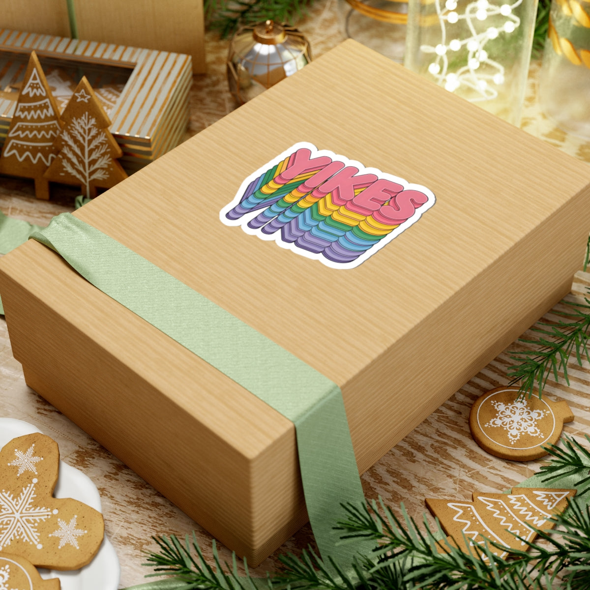 A kraft paper gift box tied with a pale green ribbon, featuring a colorful 'YIKES' sticker on top, styled with holiday cookies, pine branches, and gift decorations.