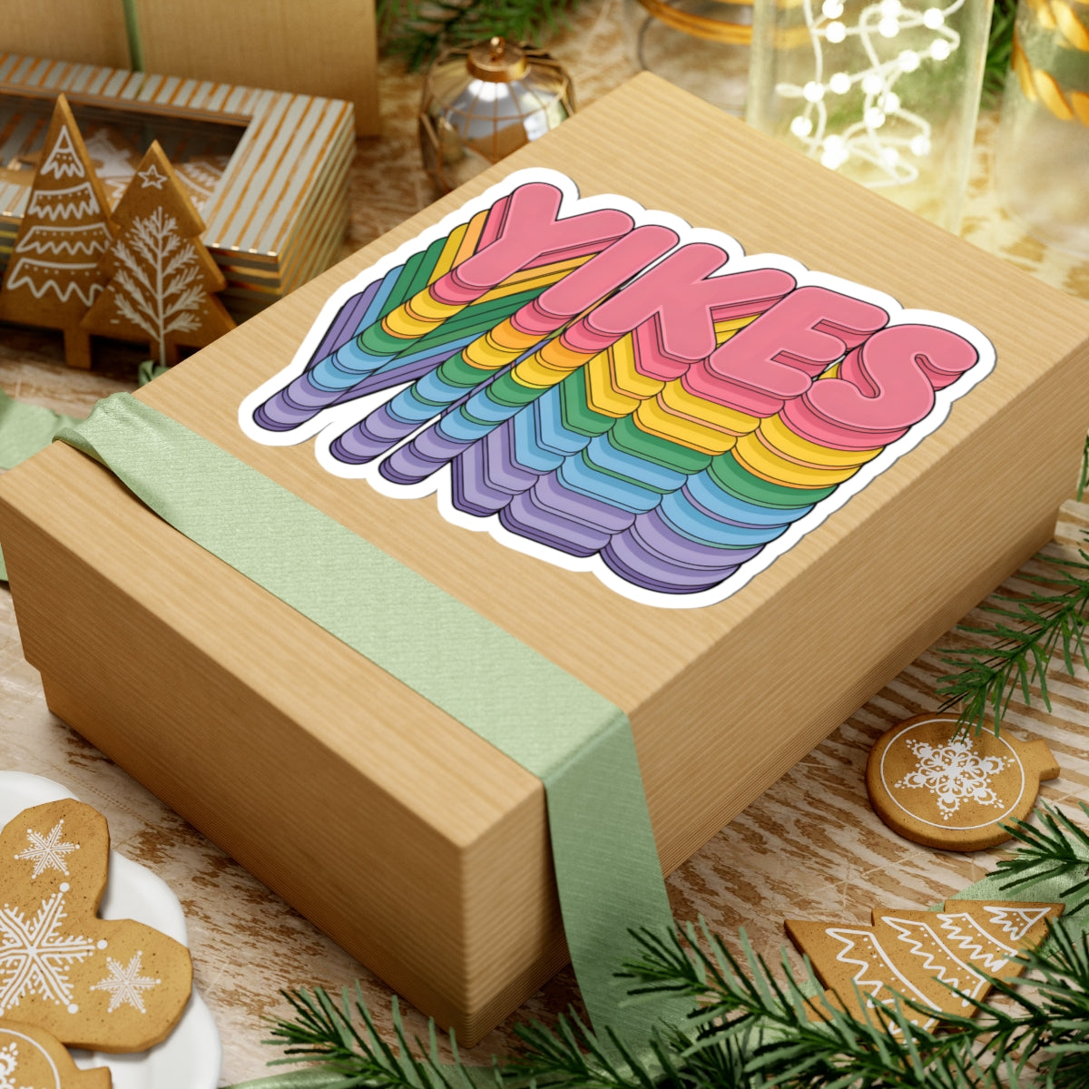 A kraft paper gift box wrapped with a pale green ribbon, featuring a large colorful sticker on top that reads “YIKES” in rainbow bubble letters. The box is styled on a rustic wooden surface with holiday cookies, evergreen branches, and warm decorative lights in the background.
