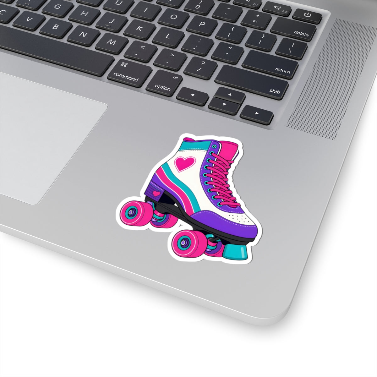 A colorful roller skate sticker applied to the silver surface of a laptop keyboard. The sticker features a white roller skate with pink, purple, and teal accents, a pink heart on the side, and pink wheels.