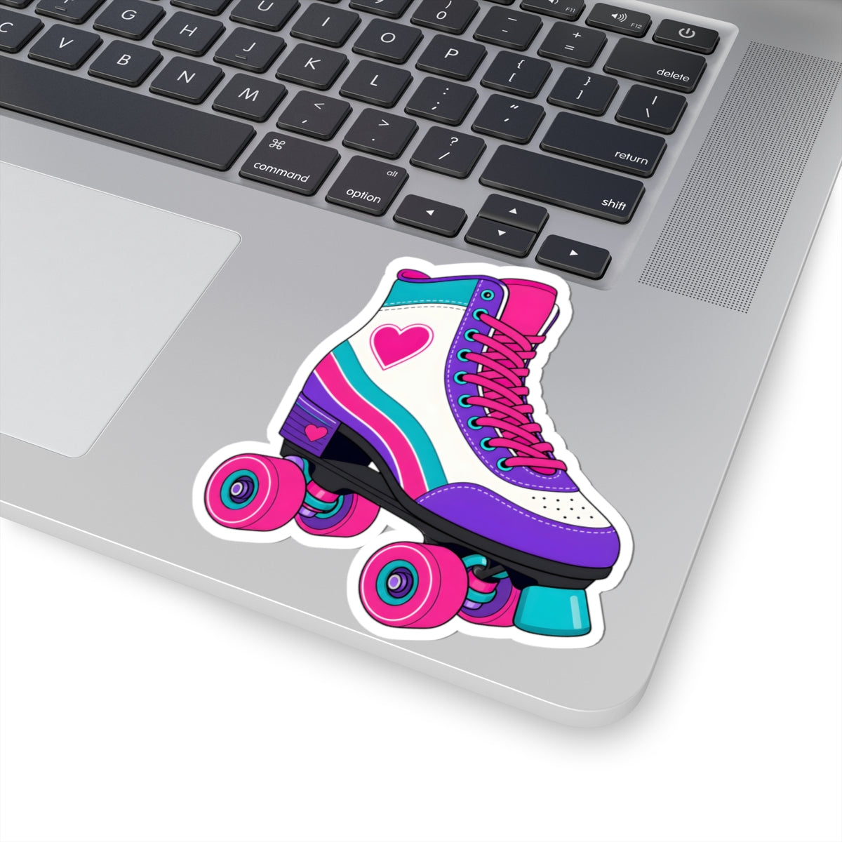 A kiss-cut vinyl sticker of a retro roller skate in pink, purple, and teal, shown applied to the silver surface of a laptop next to the keyboard.