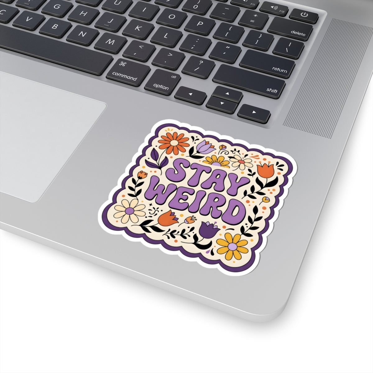 A square kiss-cut sticker with the words 'STAY WEIRD' in purple, surrounded by colorful retro flowers and doodles, shown applied to the palm rest of a silver laptop.