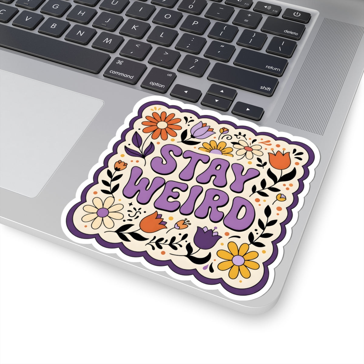 A kiss-cut vinyl sticker with the text 'STAY WEIRD' in purple, decorated with colorful flowers and black floral outlines, shown applied to the corner of a silver laptop.