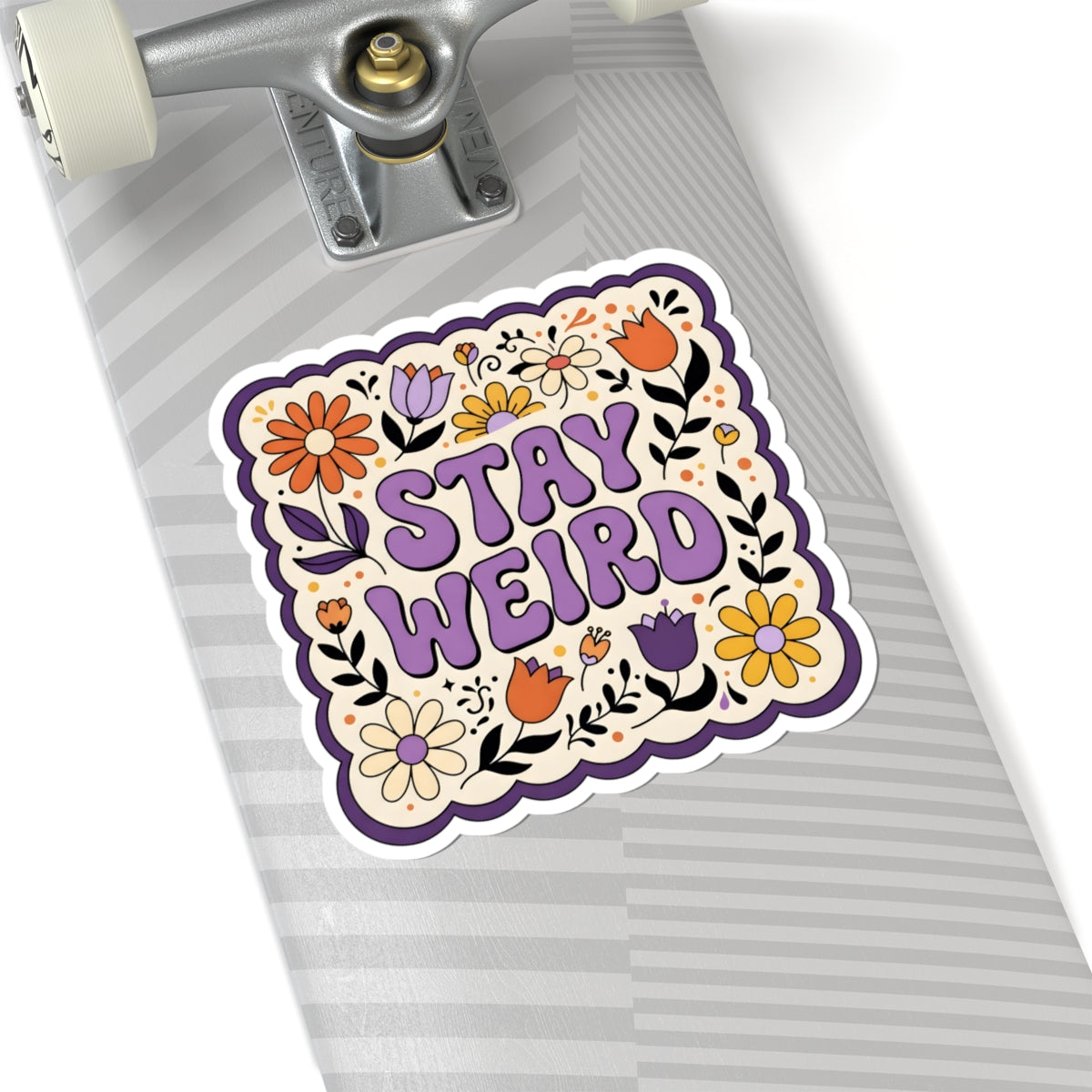 A kiss-cut sticker with the words 'STAY WEIRD' in purple, surrounded by colorful flowers and leaves, shown applied to the underside of a skateboard.