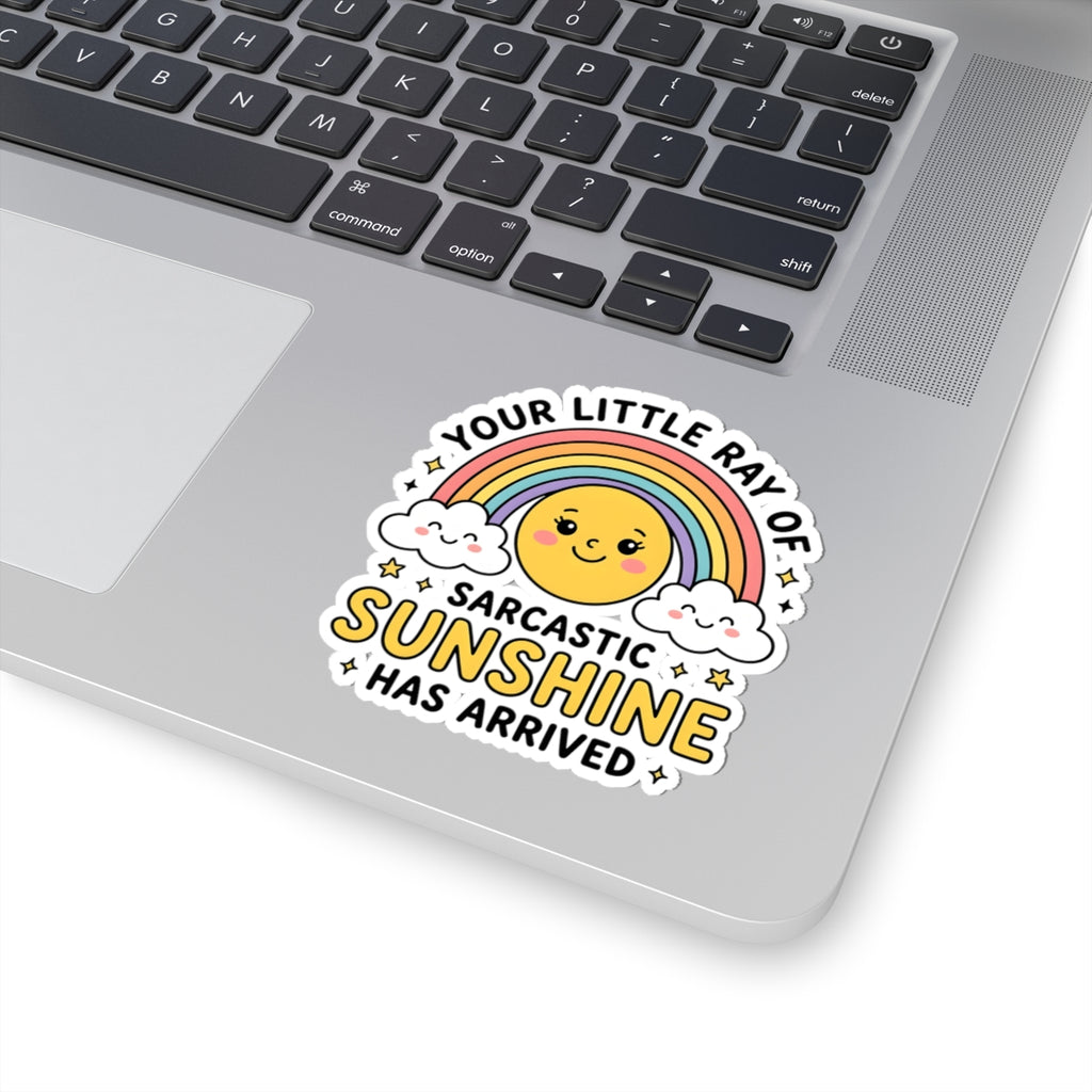 A kiss-cut vinyl sticker with a cute rainbow sun graphic and the text 'YOUR LITTLE RAY OF SARCASTIC SUNSHINE HAS ARRIVED' applied to the palm rest of a silver laptop.