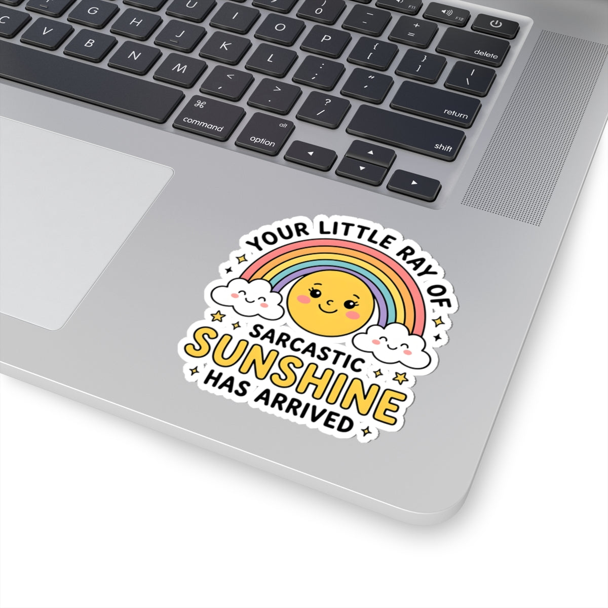 A kiss-cut vinyl sticker with a cute rainbow sun graphic and the text 'YOUR LITTLE RAY OF SARCASTIC SUNSHINE HAS ARRIVED' applied to the palm rest of a silver laptop.