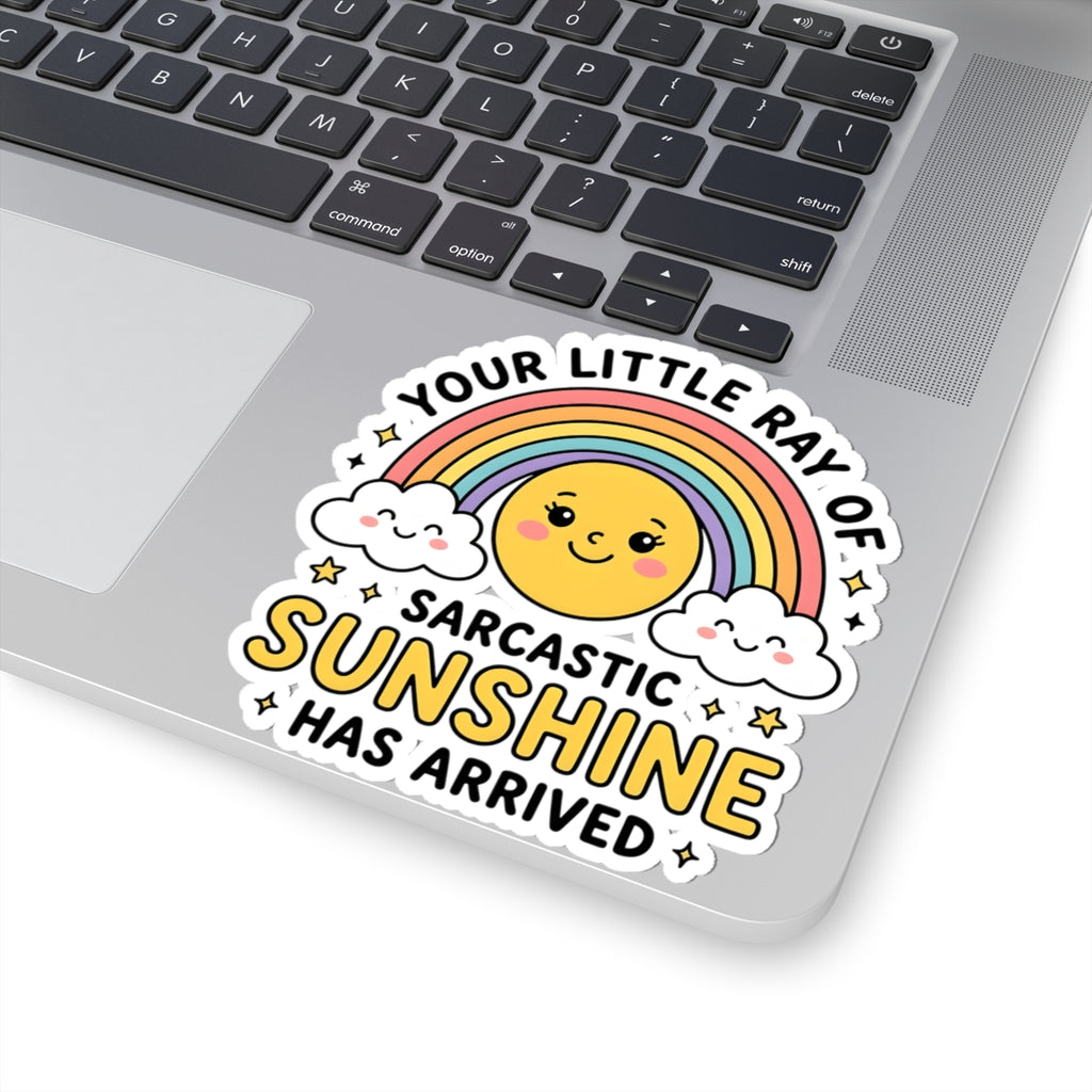 A KISS-cut vinyl sticker with a cute cartoon smiley face, rainbow, clouds, and the text 'YOUR LITTLE RAY OF SARCASTIC SUNSHINE HAS ARRIVED' applied to the lower left corner of a silver laptop.