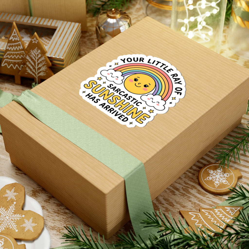 A square kraft gift box wrapped with a pale green ribbon, decorated with a die-cut sticker that reads 'YOUR LITTLE RAY OF SARCASTIC SUNSHINE HAS ARRIVED' around a smiling rainbow sun. The box is styled on a wooden surface with holiday cookies, greenery, and warm festive decor in the background.