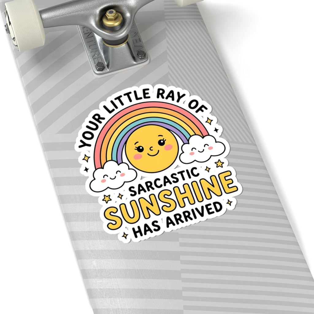 A close-up of a skateboard deck with a colorful sticker that reads 'YOUR LITTLE RAY OF SARCASTIC SUNSHINE HAS ARRIVED' around a smiling sun, rainbow, and clouds. The skateboard has a gray and white striped pattern and is shown against a white background.