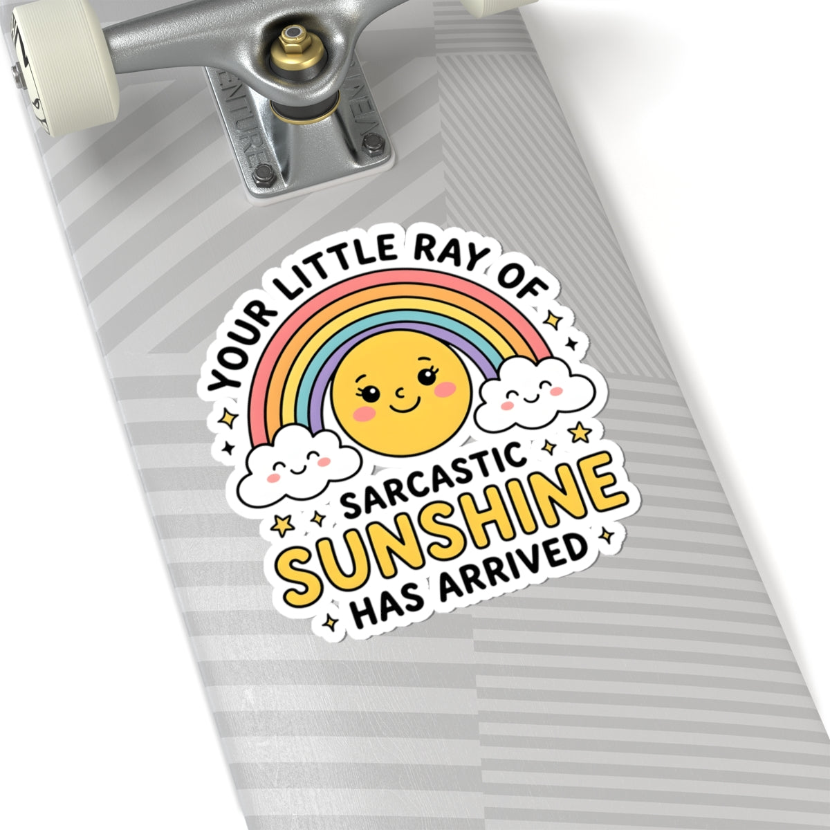 A close-up of a skateboard deck with a colorful sticker that reads 'YOUR LITTLE RAY OF SARCASTIC SUNSHINE HAS ARRIVED' around a smiling sun, rainbow, and clouds. The skateboard has a gray and white striped pattern and is shown against a white background.