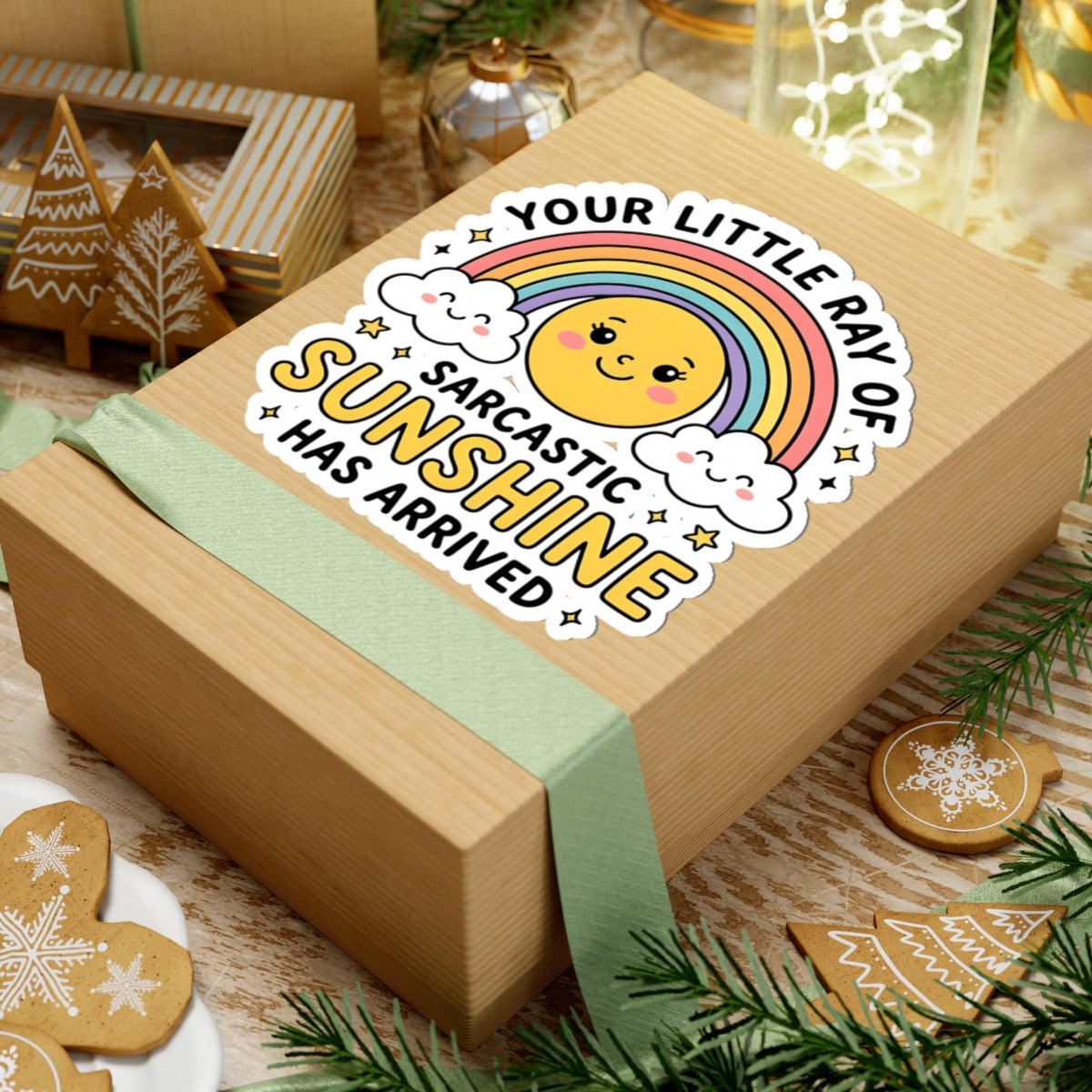 A kraft paper gift box wrapped with a pale green ribbon, shown in a styled holiday setting. On top of the box is a kiss-cut sticker featuring a smiling yellow sun, clouds, and a rainbow with the text 'YOUR LITTLE RAY OF SARCASTIC SUNSHINE HAS ARRIVED'.