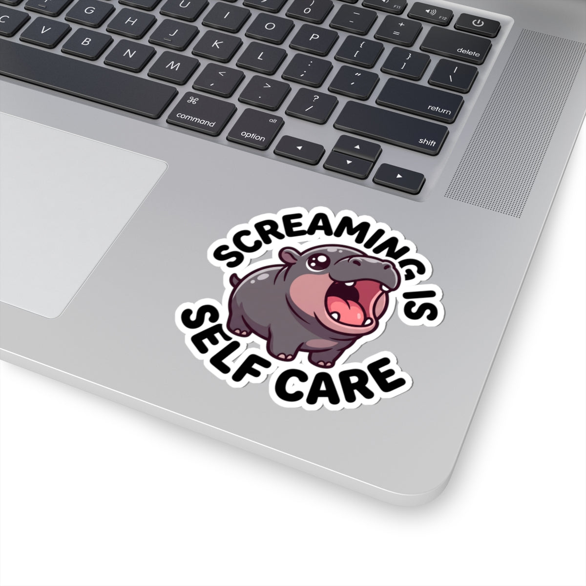 A kiss-cut vinyl sticker featuring a cartoon hippopotamus and the text 'SCREAMING IS SELF CARE' applied to the palm rest of a silver laptop.