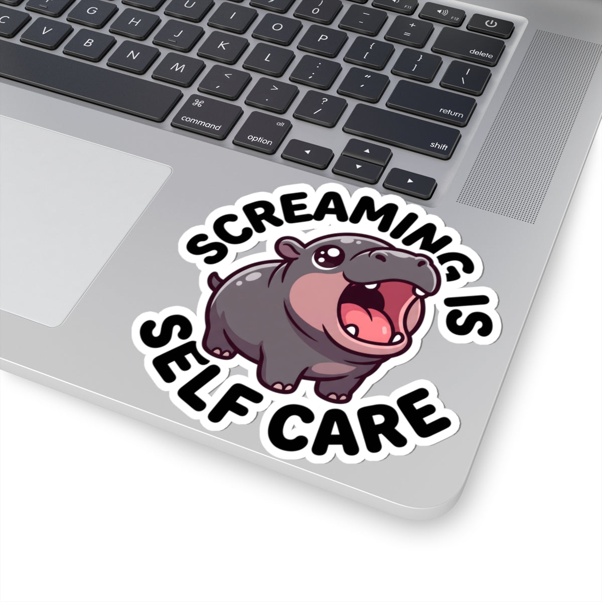 A kiss-cut vinyl sticker applied to the corner of a silver laptop. The sticker features a cartoon hippopotamus with its mouth wide open, surrounded by the text 'SCREAMING IS SELF CARE' in bold black lettering with a white outline.