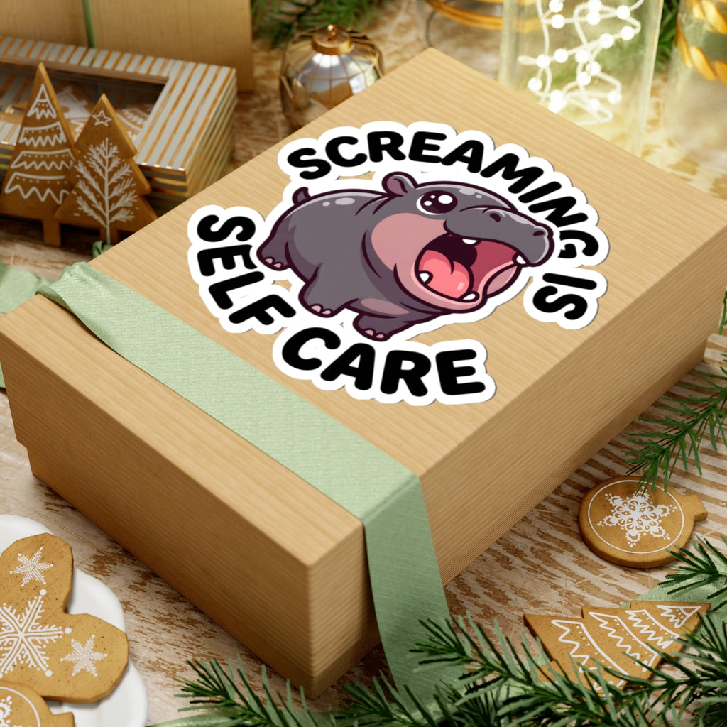 A square kraft gift box wrapped with a pale green ribbon, topped with a kiss-cut sticker featuring a cartoon hippo and the text “SCREAMING IS SELF CARE.” The box is styled on a wooden surface with holiday decorations including gingerbread cookies, evergreen branches, and small boxes.