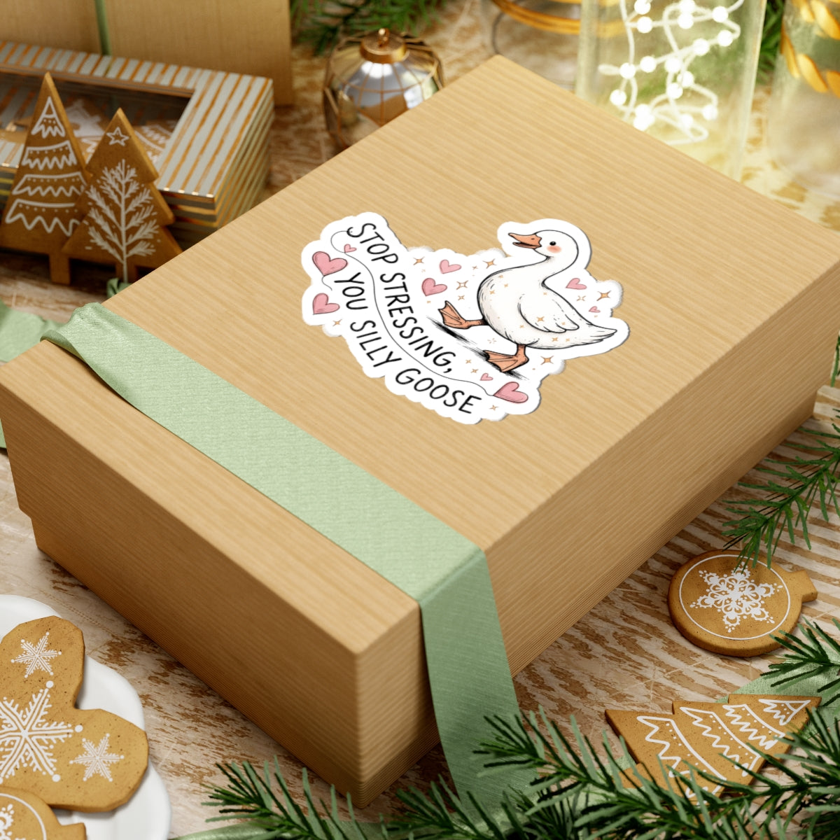 A square kraft paper gift box wrapped with a pale green ribbon, featuring a kiss-cut sticker of a white goose with the text 'STOP STRESSING, YOU SILLY GOOSE' and pink hearts. The box is styled on a rustic wooden surface with gingerbread cookies, pine branches, and holiday decorations.