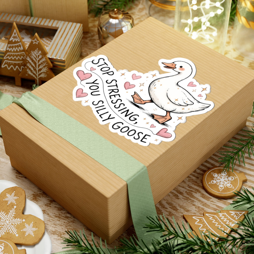 A square kraft gift box wrapped with a pale green ribbon, decorated with a kiss-cut sticker of a white goose surrounded by pink hearts and the text 'STOP STRESSING, YOU SILLY GOOSE.' The box is styled on a wooden surface with holiday decorations including gingerbread cookies, evergreen branches, and small ornaments.