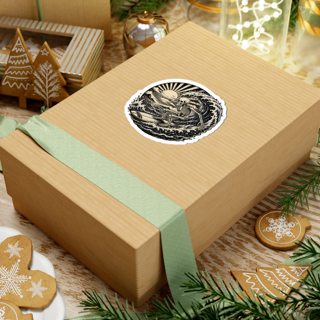 A square kraft paper gift box wrapped with a pale green ribbon, topped with a round black-and-cream sticker featuring a retro surfing skeleton, styled on a tabletop with holiday cookies, pine branches, and other gift items.