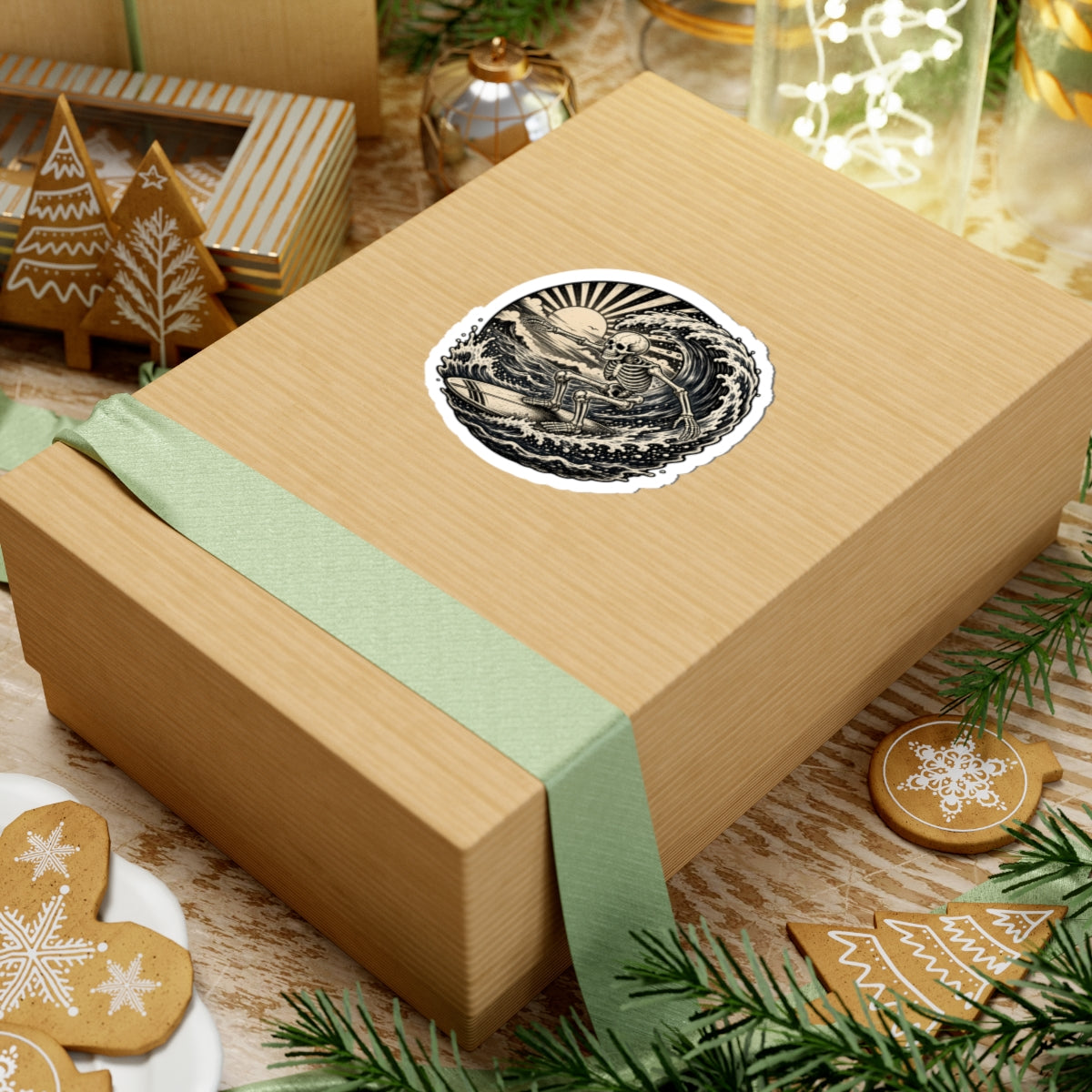 A square kraft paper gift box wrapped with a pale green ribbon, topped with a round black-and-cream sticker featuring a retro surfing skeleton, styled on a tabletop with holiday cookies, pine branches, and other gift items.