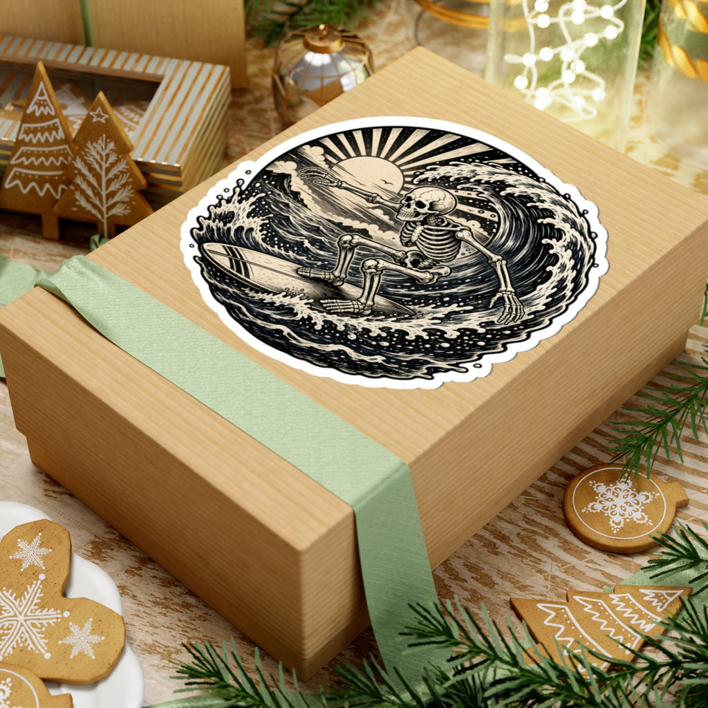 A round sticker with a black and beige skeleton surfing graphic is shown on top of a kraft gift box wrapped with a pale green ribbon. The scene is styled with holiday decorations, including gingerbread cookies, pine branches, and a small wooden ornament.