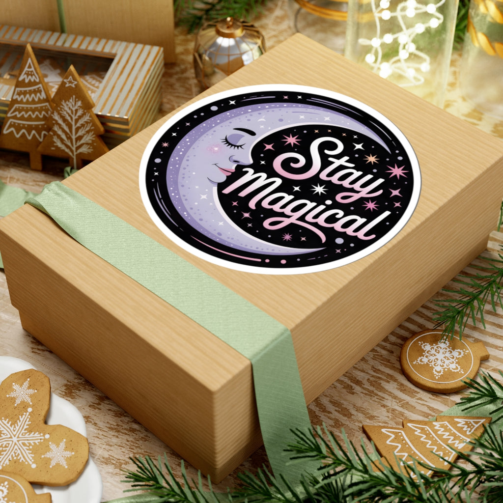 A round sticker with the words 'Stay Magical' in pink script over a purple crescent moon and starry night design, shown placed on top of a kraft paper gift box wrapped with a pale green ribbon. The scene is styled with holiday cookies, pine branches, and warm background decor.