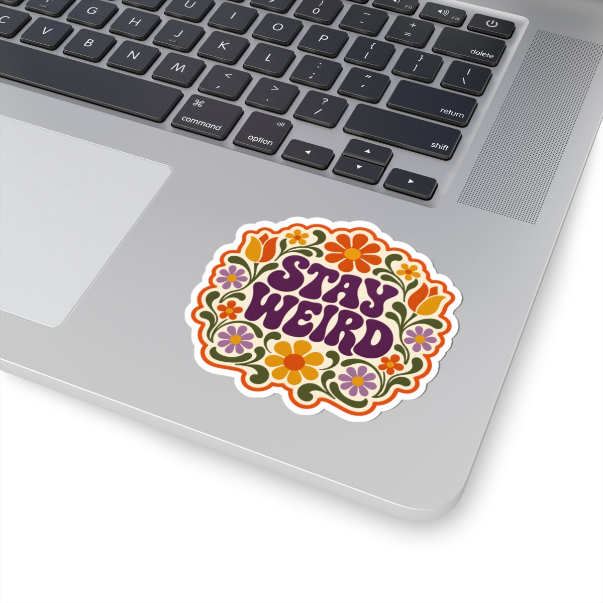 A kiss-cut vinyl sticker with the phrase 'STAY WEIRD' in purple retro lettering, surrounded by a colorful retro floral pattern, shown applied to the silver palm rest of a laptop.