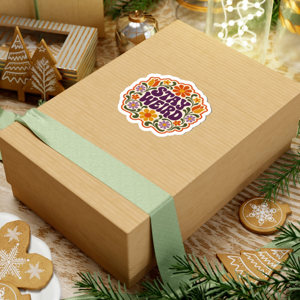 Gift box with a green ribbon and a retro "STAY WEIRD" floral sticker on a festive background with cookies and lights.
