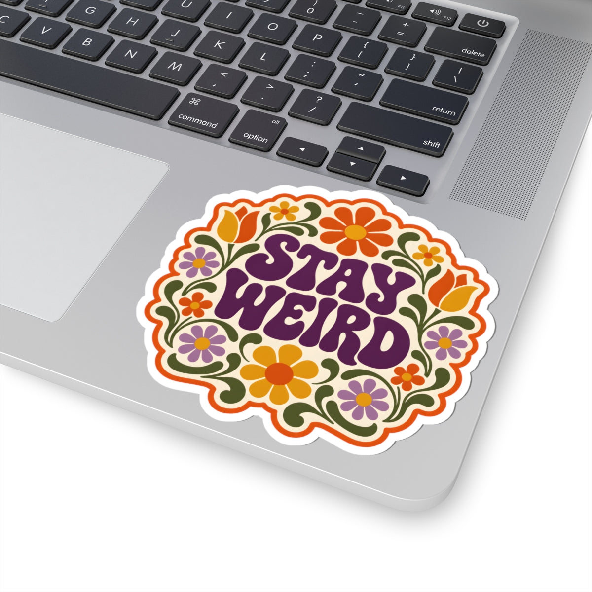 A kiss-cut vinyl sticker with the text 'STAY WEIRD' in purple retro lettering, surrounded by colorful retro flowers and green vines, shown applied to the corner of a silver laptop.