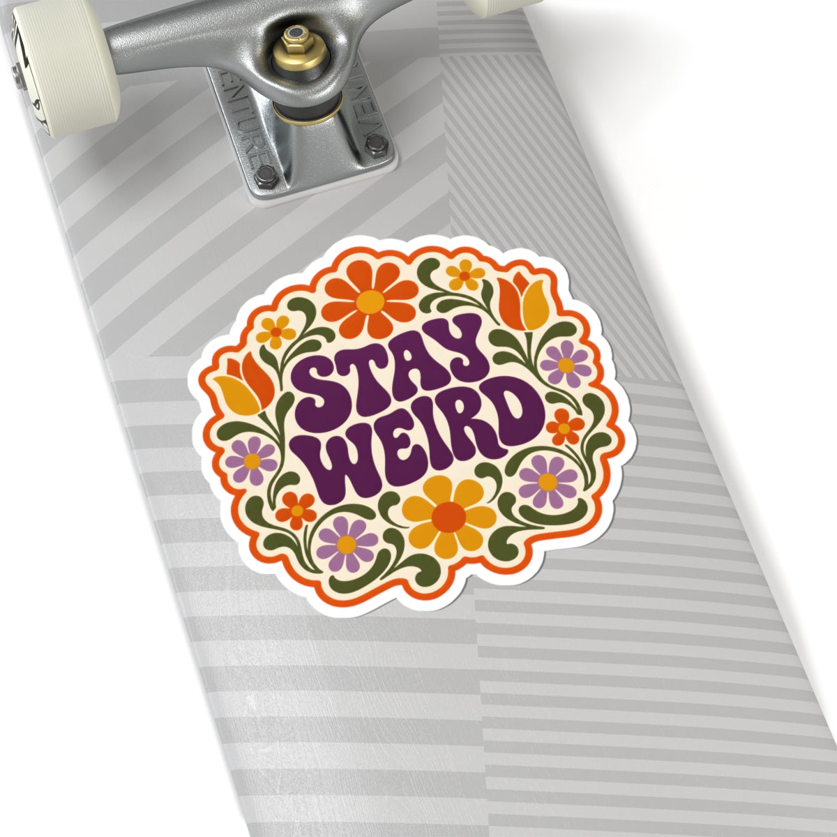 A kiss-cut sticker with the words 'STAY WEIRD' in purple retro lettering, surrounded by colorful retro flowers and green leaves, shown applied to the deck of a skateboard.