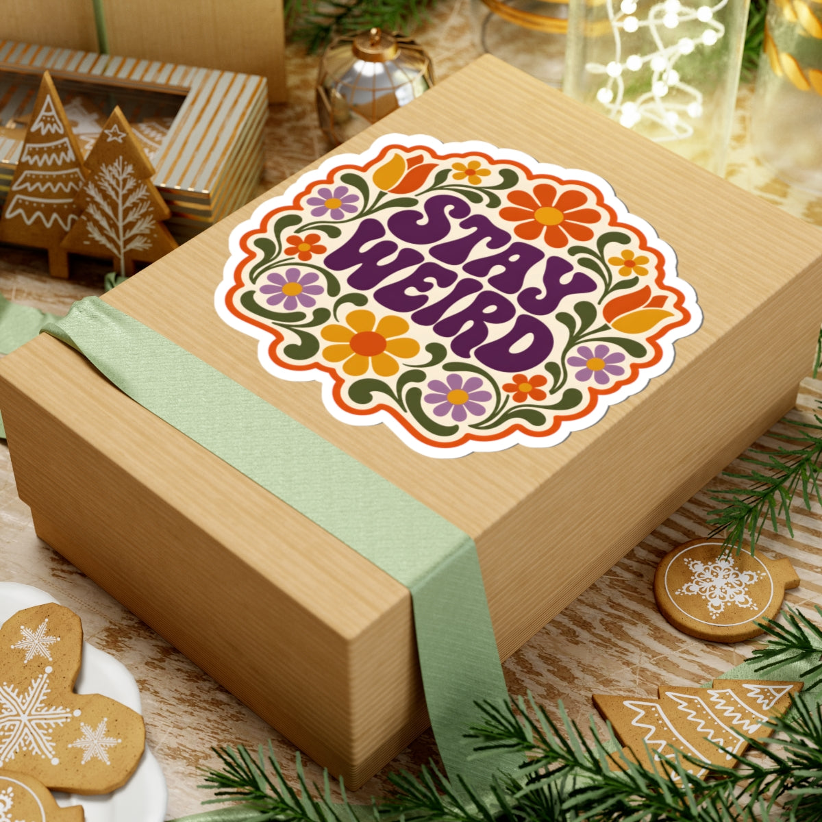 A kraft paper gift box wrapped with a pale green ribbon, topped with a colorful retro-style sticker that reads “STAY WEIRD.” The box is styled on a wooden surface with holiday cookies, evergreen branches, and warm ambient decor in the background.