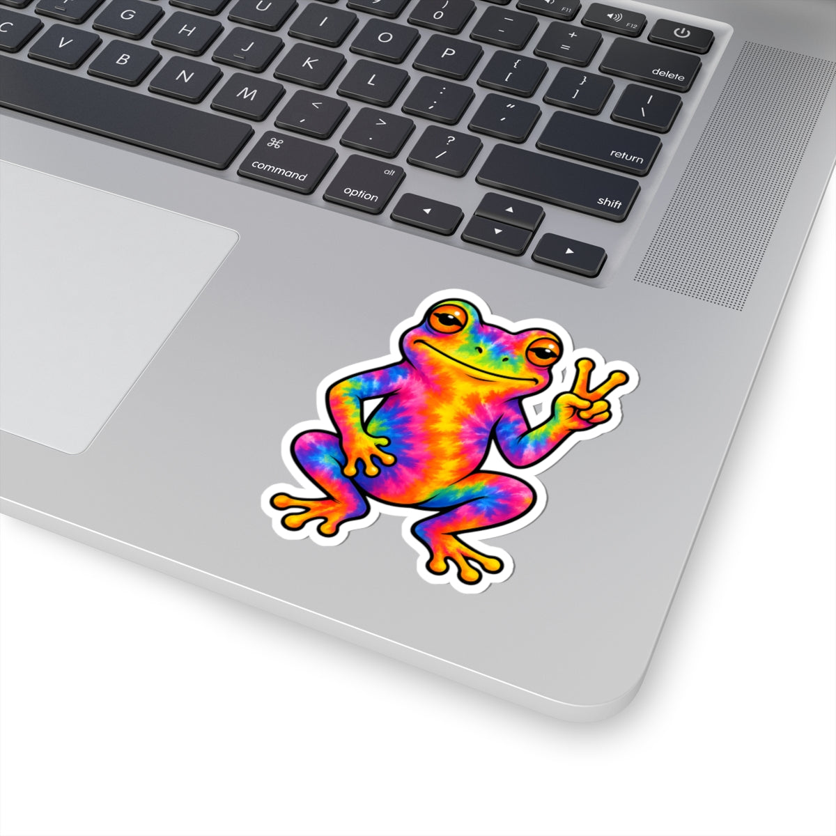 A kiss-cut vinyl sticker of a colorful tie-dye frog making a peace sign, shown applied to the palm rest of a silver laptop.