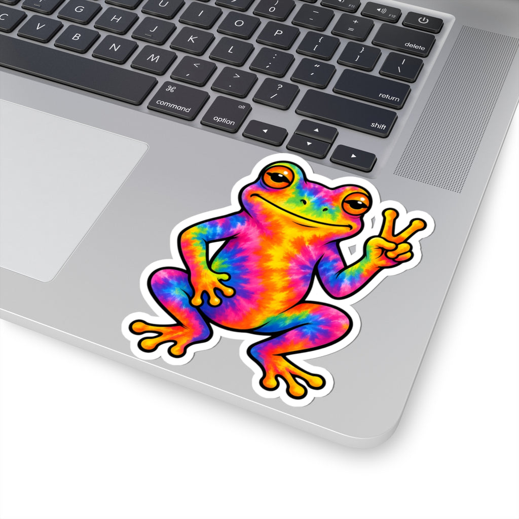 A kiss-cut vinyl sticker of a colorful tie-dye frog making a peace sign, shown applied to the silver surface of a laptop keyboard.