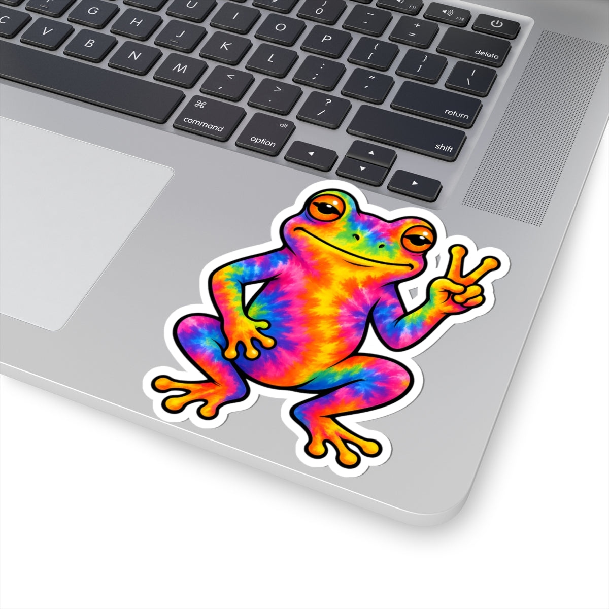 A kiss-cut vinyl sticker of a colorful tie-dye frog making a peace sign, shown applied to the silver surface of a laptop keyboard.