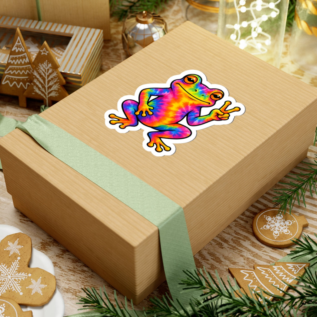 A kraft paper gift box wrapped with a pale green ribbon, featuring a colorful rainbow frog sticker on top, styled with holiday cookies, pine branches, and warm background lights.