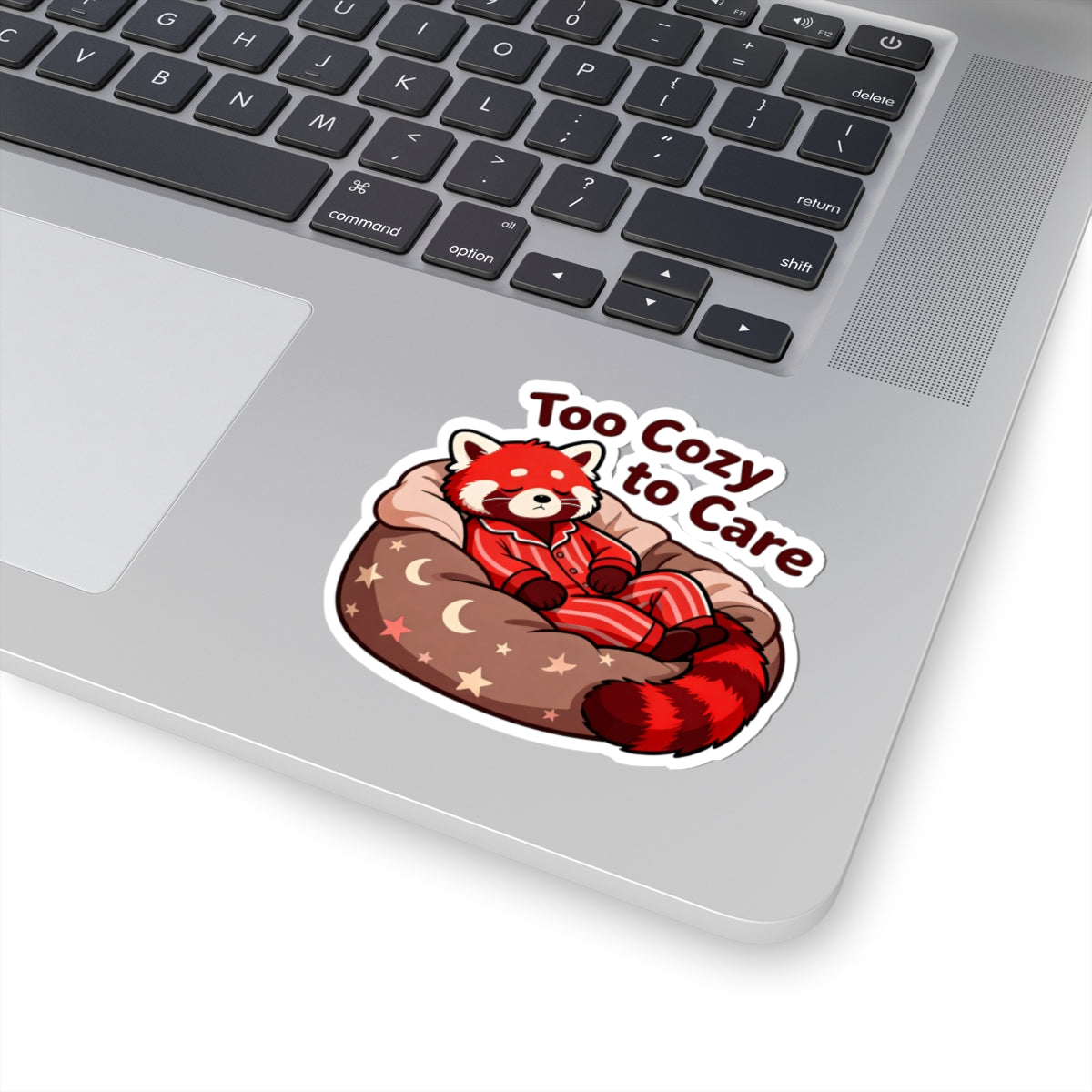 A kiss-cut vinyl sticker featuring a cartoon red panda in a cozy bed with the text 'Too Cozy to Care', shown applied to the silver surface of a laptop keyboard.
