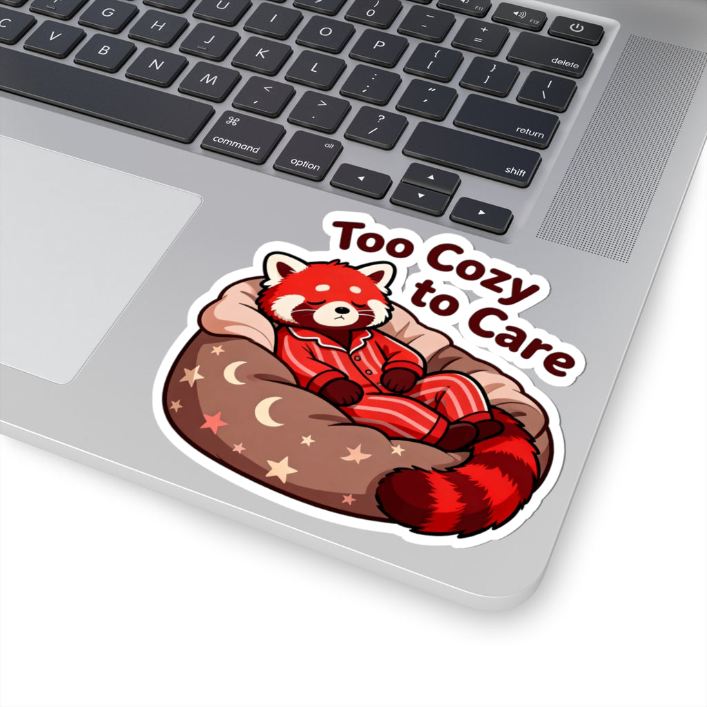 A kiss-cut vinyl sticker featuring a red panda in red pajamas lounging on a brown beanbag with stars and moons, next to the text 'Too Cozy to Care' in dark red. The sticker is shown applied to the silver surface of a laptop keyboard.
