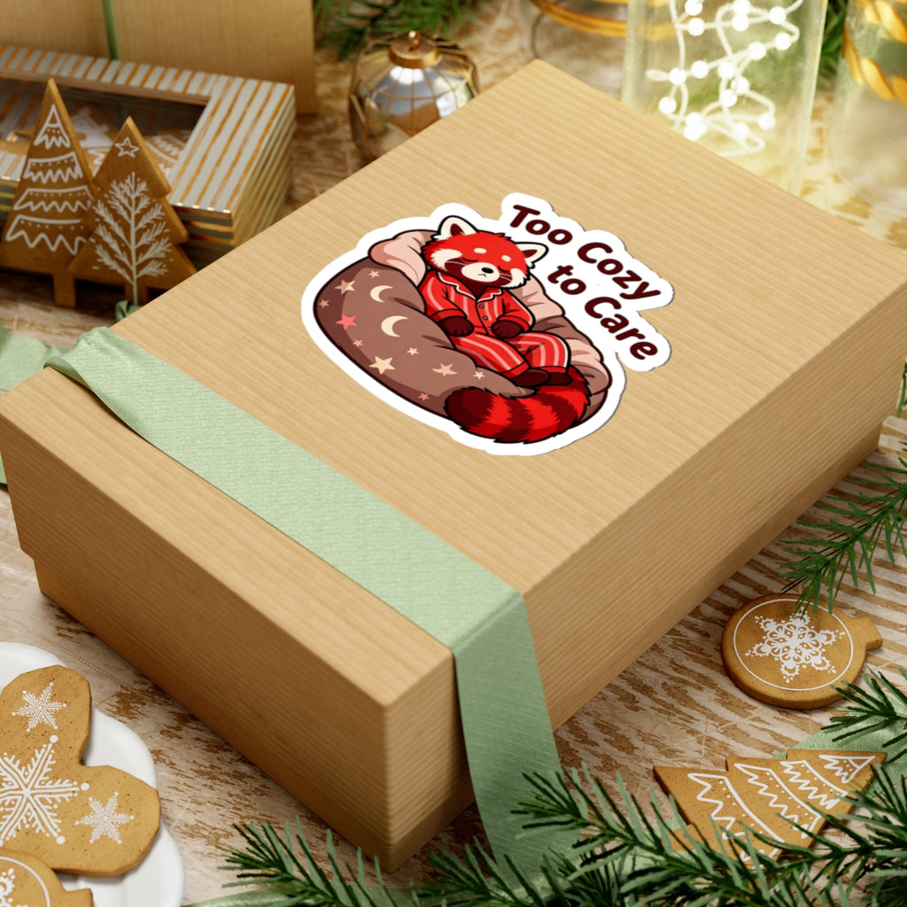 A light brown kraft gift box wrapped with a pale green ribbon, featuring a kiss-cut sticker of a red panda sleeping on a red bean bag with the text 'Too Cozy to Care'. The box is styled on a wooden surface with Christmas cookies, evergreen branches, and holiday decorations.