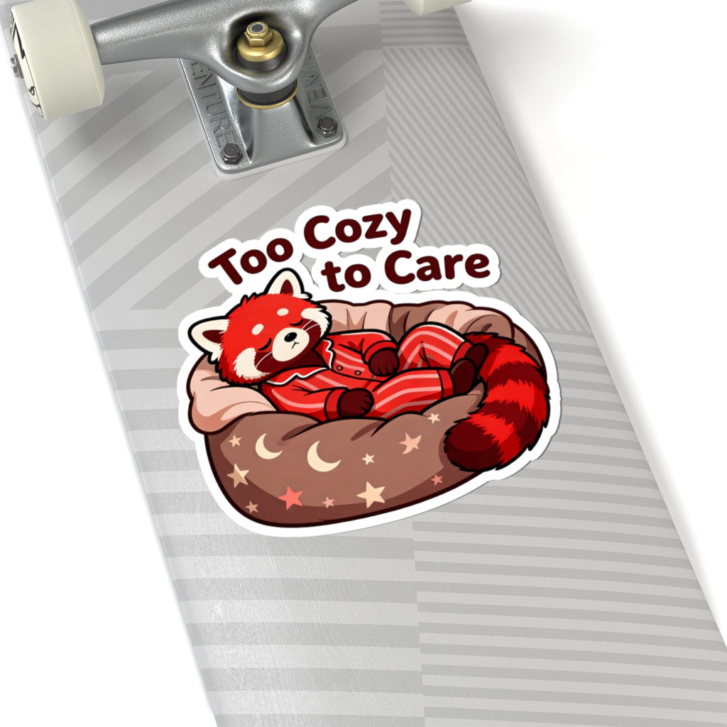 A kiss-cut sticker featuring a red panda sleeping in a cozy bed with the text 'Too Cozy to Care' on a gray striped skateboard deck.