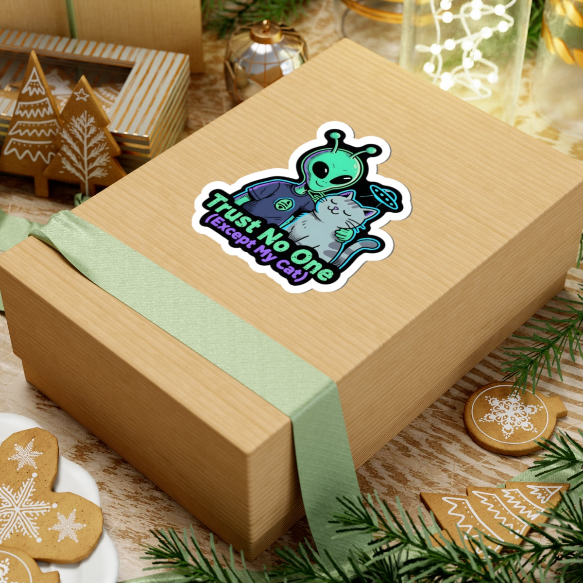 A kraft paper gift box wrapped with a pale green ribbon, topped with a kiss-cut sticker of an alien with a cat and text 'trust no one, except my cat'. The box is styled on a rustic wooden table with Christmas cookies, pine branches, and holiday decorations.