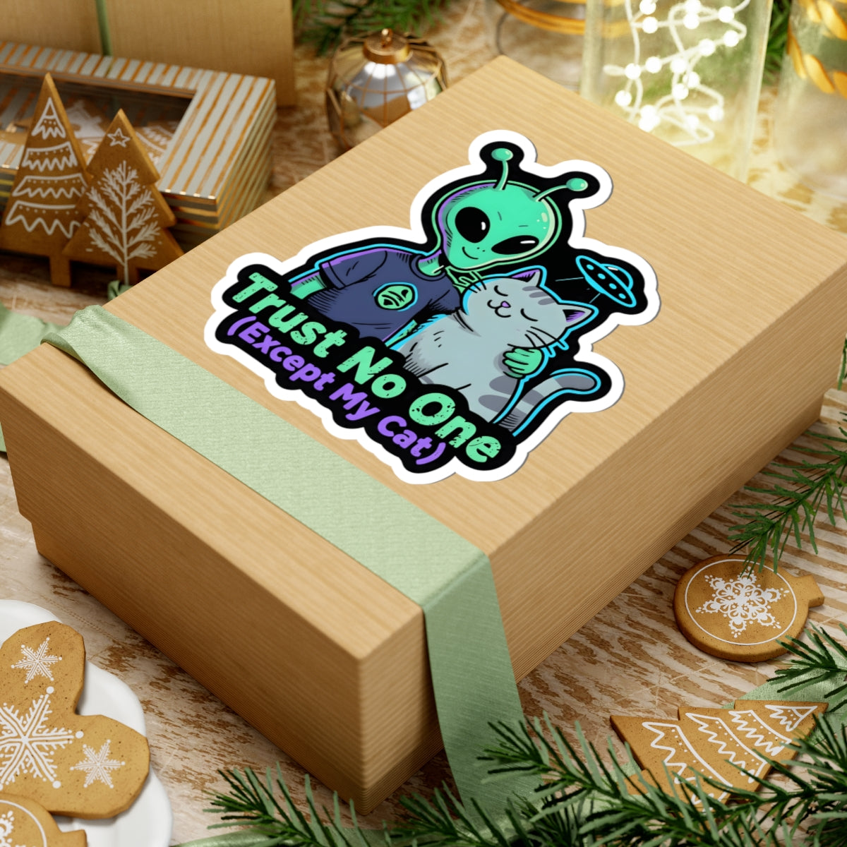 A square kraft paper gift box wrapped with a pale green ribbon, featuring a colorful cartoon sticker of a green alien holding a gray cat with the text 'Trust No One (Except My cat)'. The box is styled on a wooden surface with holiday decorations including gingerbread cookies, evergreen branches, and a small lantern.