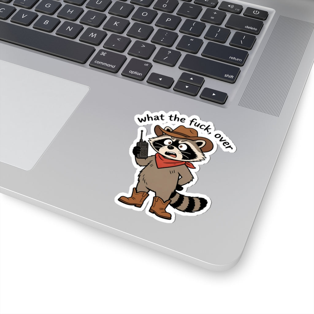 A kiss-cut vinyl sticker of a cartoon raccoon dressed as a cowboy with the text 'what the fuck over' applied to the palm rest of a silver laptop.