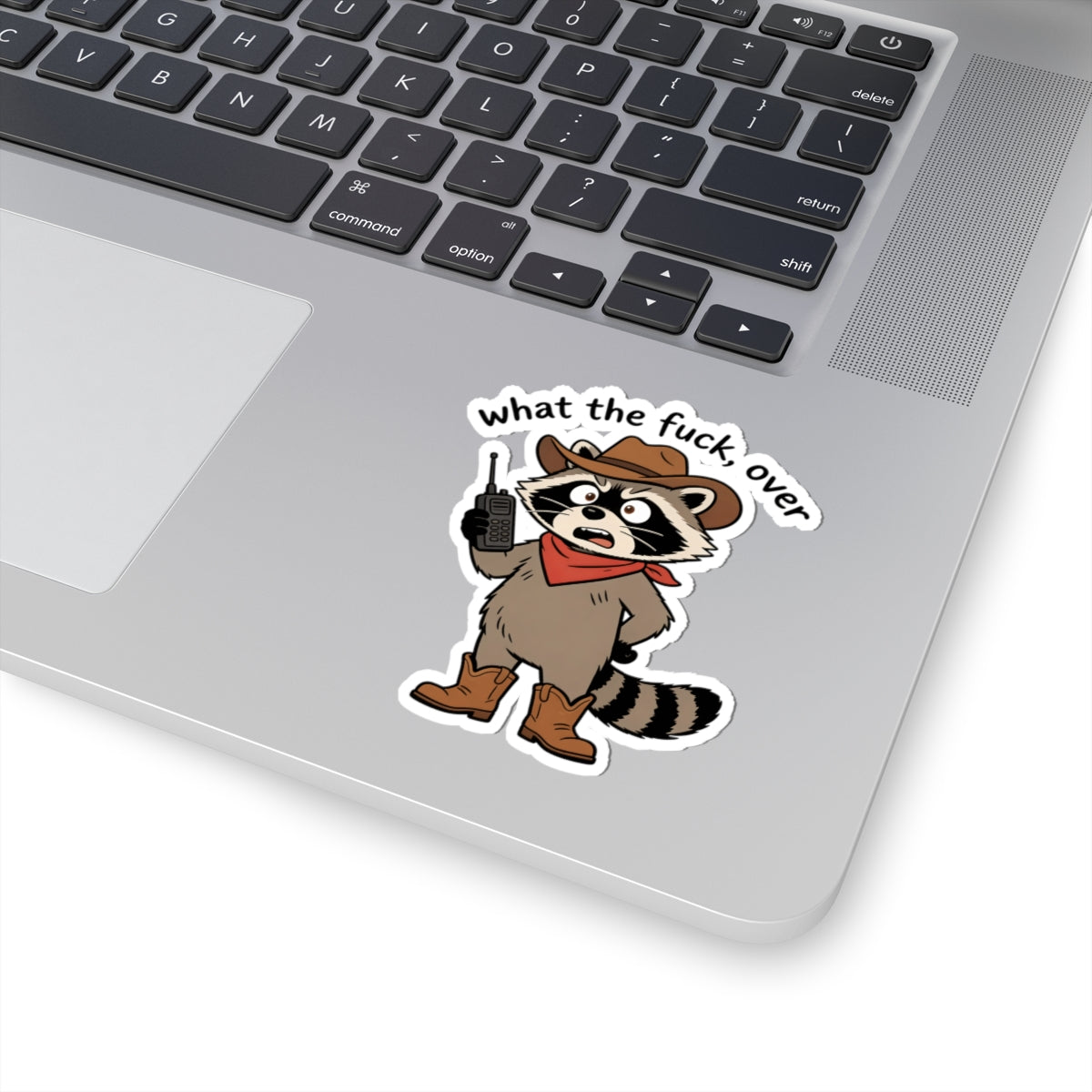 A kiss-cut vinyl sticker of a cartoon raccoon dressed as a cowboy with the text 'what the fuck over' applied to the palm rest of a silver laptop.