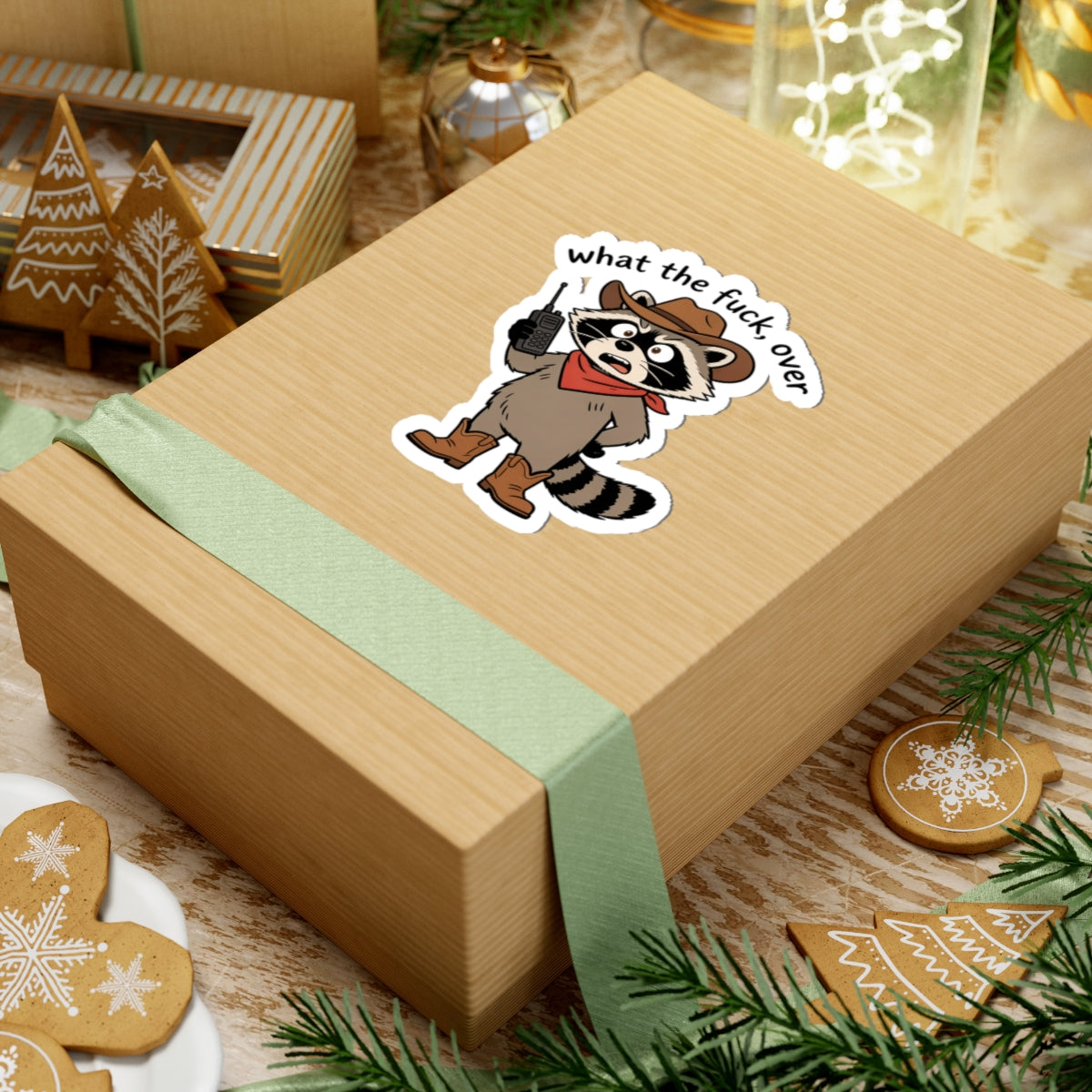 A light wood gift box tied with a pale green ribbon, featuring a kiss-cut sticker of a raccoon wearing a cowboy hat and red bandana with the text 'what the fuck over'. The box is styled on a rustic wooden surface with Christmas cookies, evergreen branches, and holiday decorations.