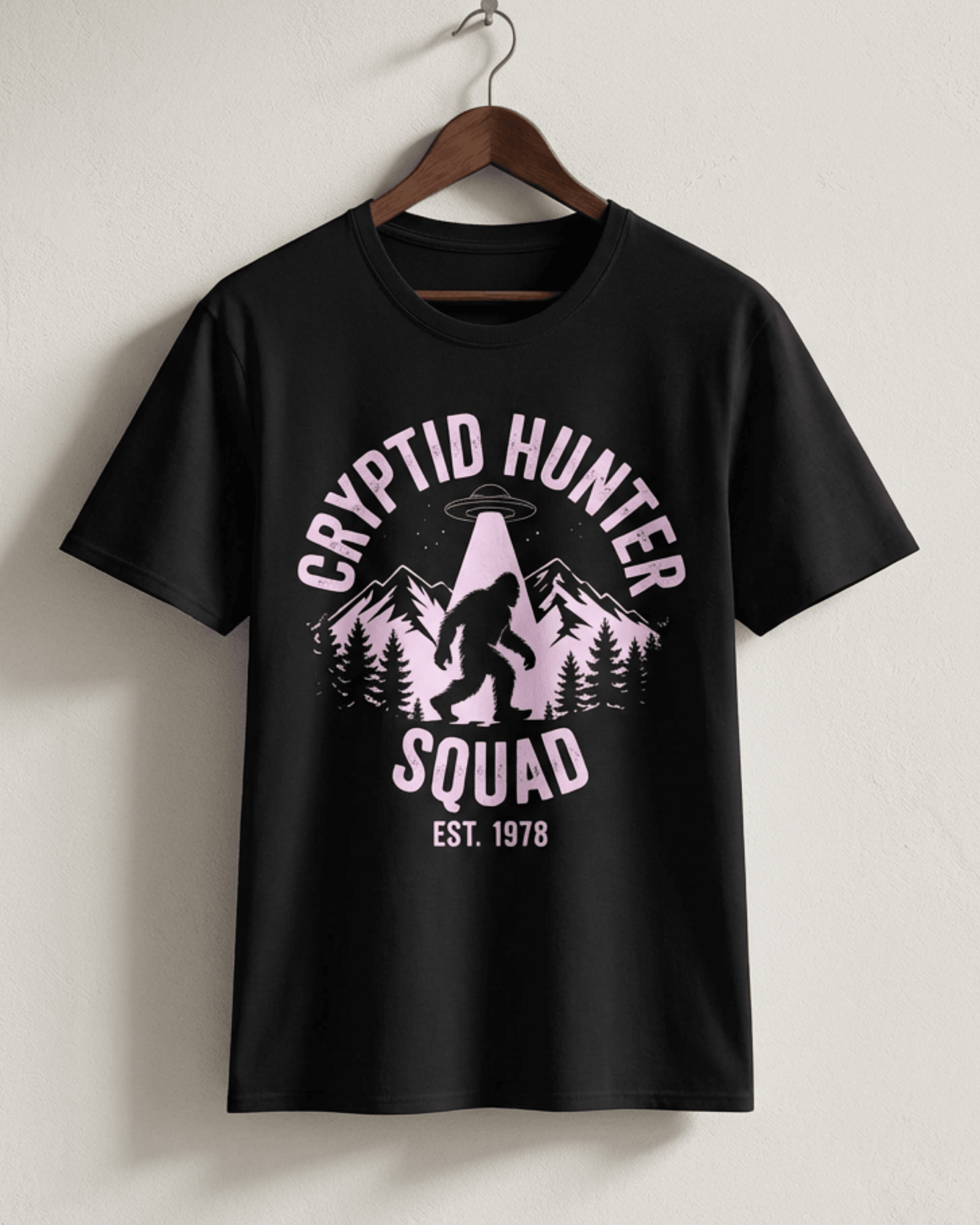 Hanger iew of the black Bigfoot Cryptid Hunter T-Shirt. The detailed pink graphic shows Sasquatch in a mountain forest scene under a UFO, with the text 'Cryptid Hunter Squad Est. 1978'. This unisex graphic tee is perfect for any cryptid enthusiast.