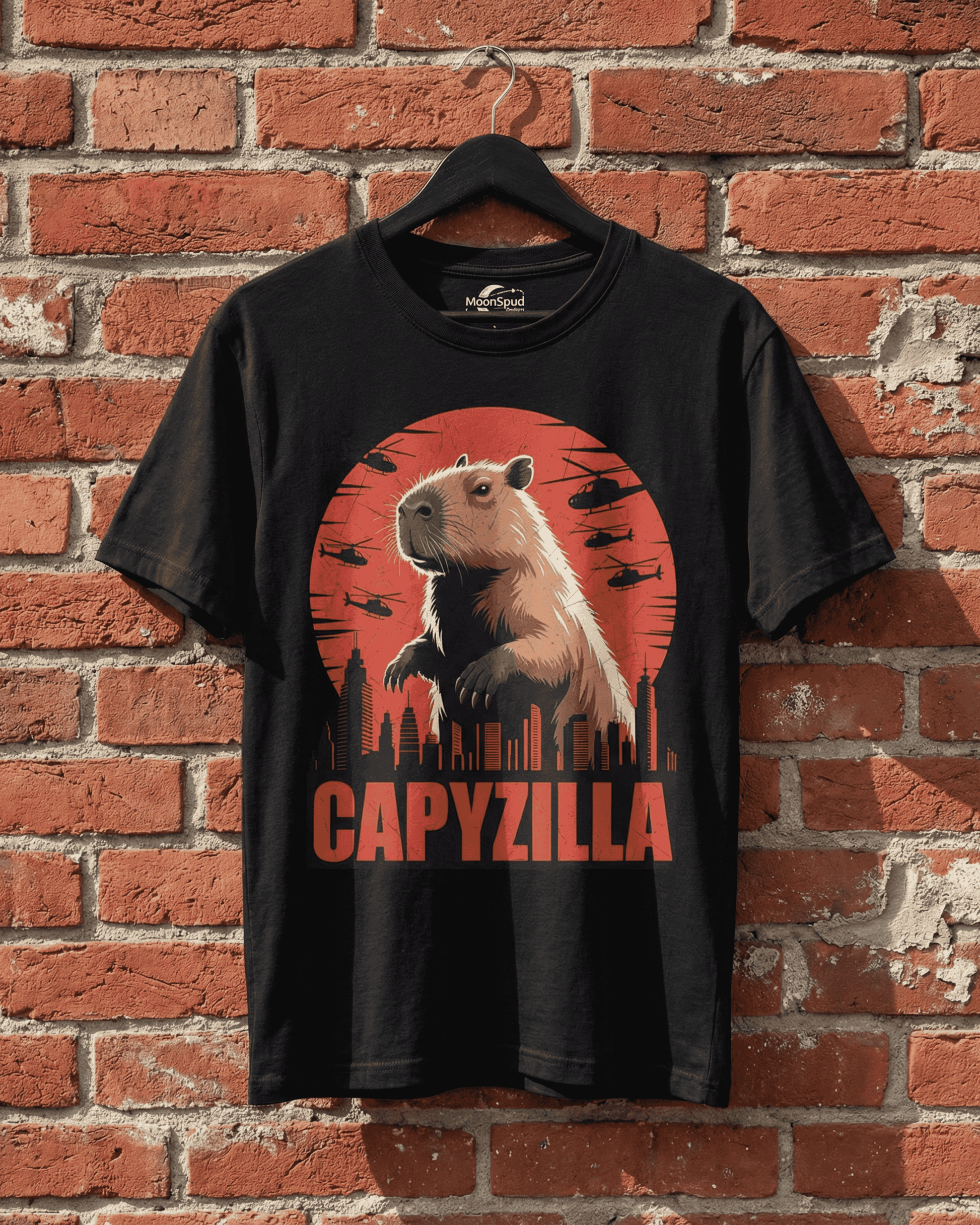 A black Funny Capybara Monster T-Shirt displayed on a hanger against a textured red brick wall. The central 'CAPYZILLA' Godzilla Parody graphic is clearly visible, illustrating a colossal capybara overlooking a city at sunset with circling helicopters. This image provides a clear view of the print and the overall design of this distinctive graphic tee.